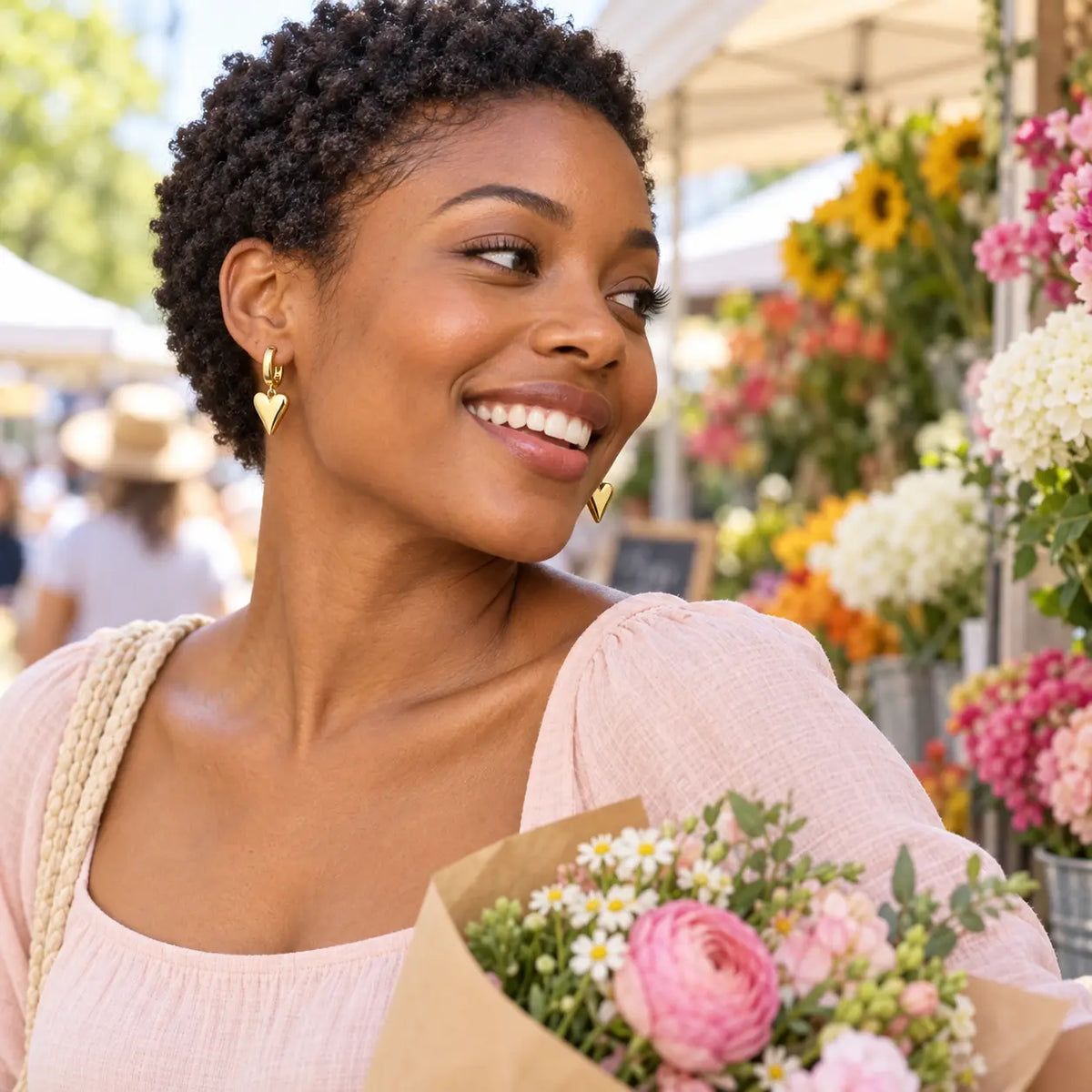 A woman with short curly black hair smiles while wearing gold heart-shaped dangle earrings, a light pink top, and holding a bouquet of pink and white flowers at an outdoor flower market.