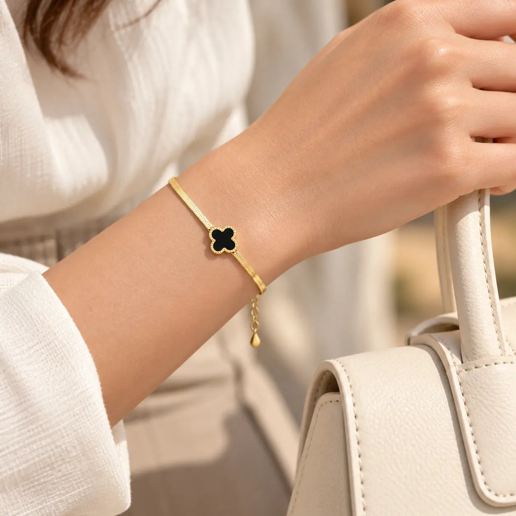 A gold-tone chain bracelet with a black enamel four-leaf clover charm, worn on a woman's wrist next to a white handbag.