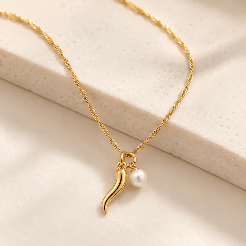 Close-up of a gold Italian horn and white pearl charm necklace with a delicate Singapore chain displayed on a soft ivory stone surface.