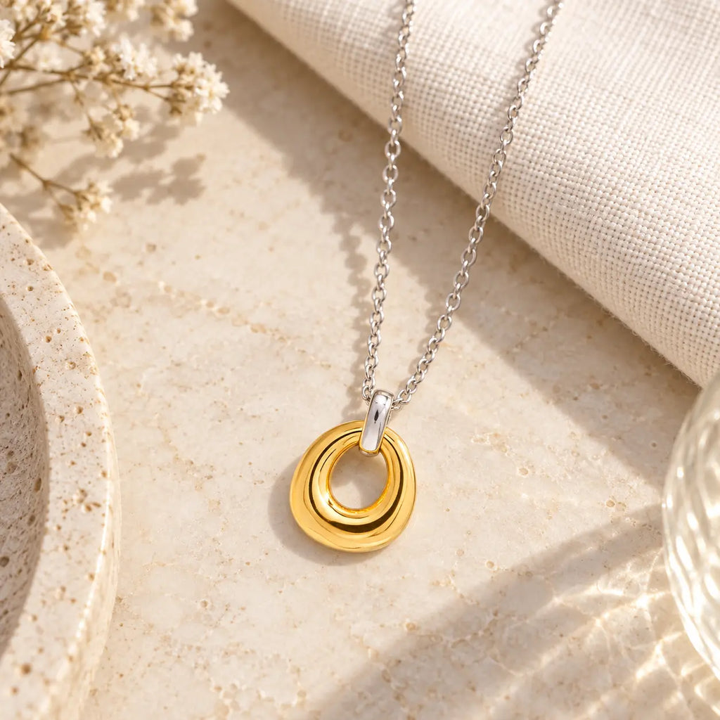 A two-tone silver and gold necklace with a double-ring pendant, styled on a textured beige stone surface with a linen cloth and dried flowers.