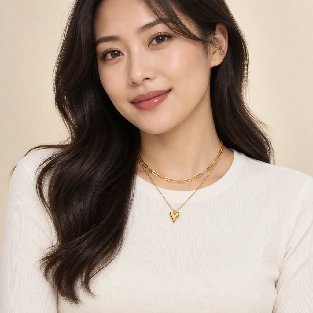 A woman with long dark wavy hair wearing a white ribbed top and layered gold necklaces, including a heart-shaped pendant, against a soft beige background.