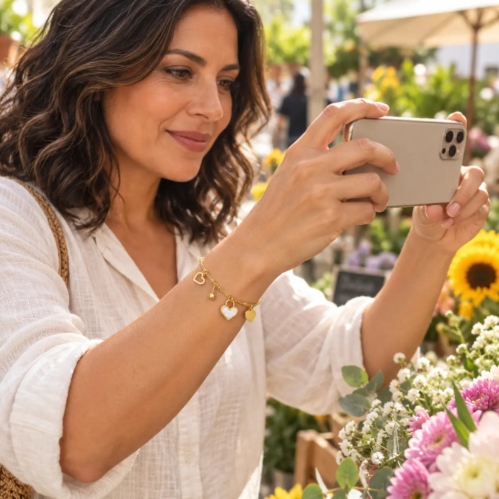 A woman in a white linen shirt takes a photo with her smartphone at an outdoor flower market, wearing a delicate gold chain bracelet with a white heart charm.