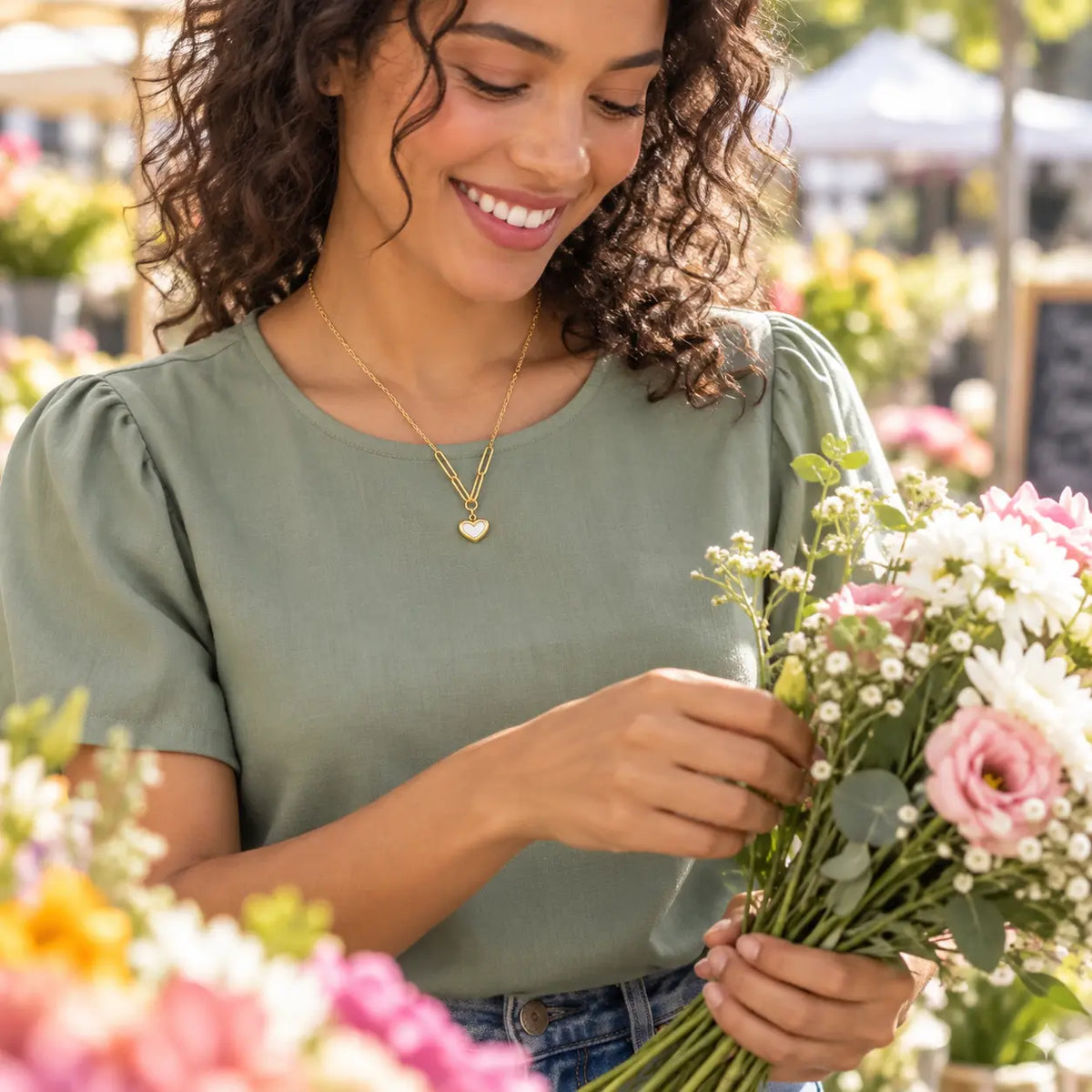 A woman with curly brown hair smiling while holding a bouquet of pink and white flowers. She is wearing an olive green short-sleeve blouse and a gold heart necklace, set against a blurred outdoor flower market background.