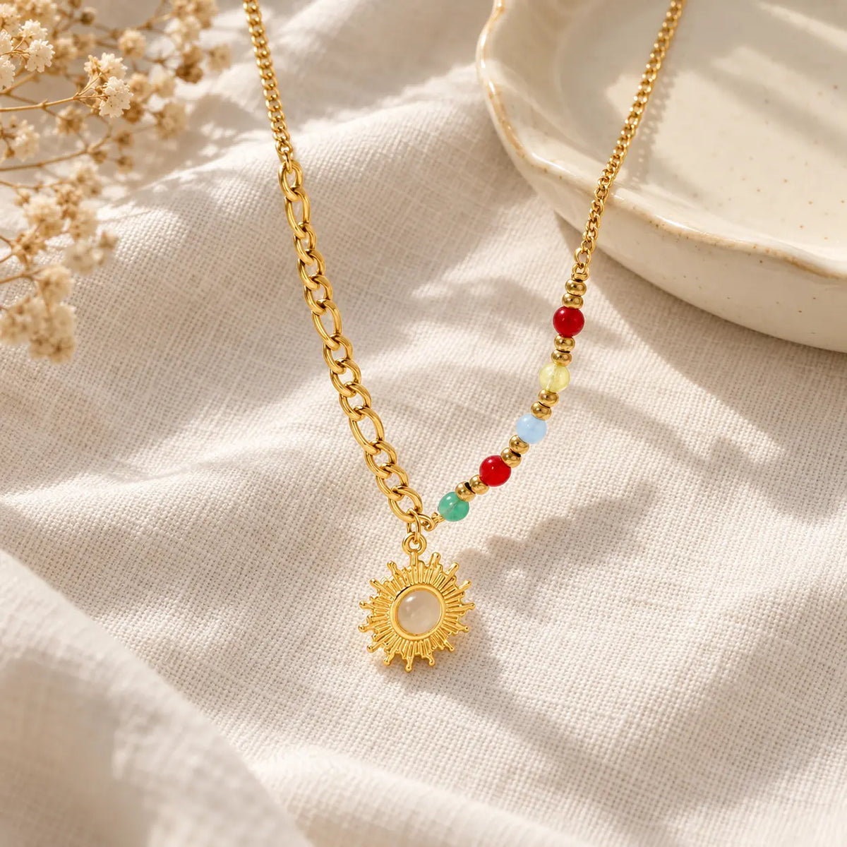 A gold chain necklace with a sun pendant and small multicolor beads, displayed on a textured cream linen fabric with dried flowers and a ceramic dish in the background.