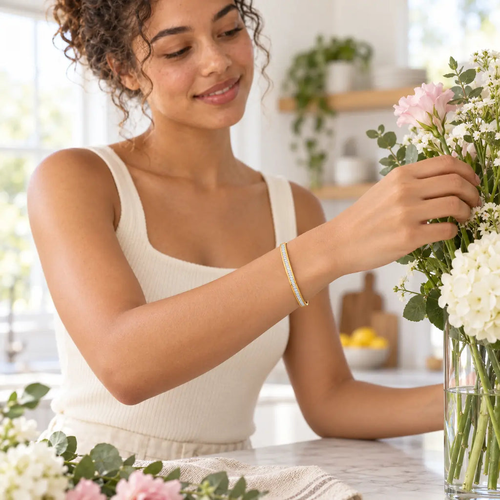 A woman in a cream ribbed tank top adjusts a bouquet of pink and white flowers in a glass vase, wearing a delicate gold chain bracelet on her wrist in a bright kitchen setting.