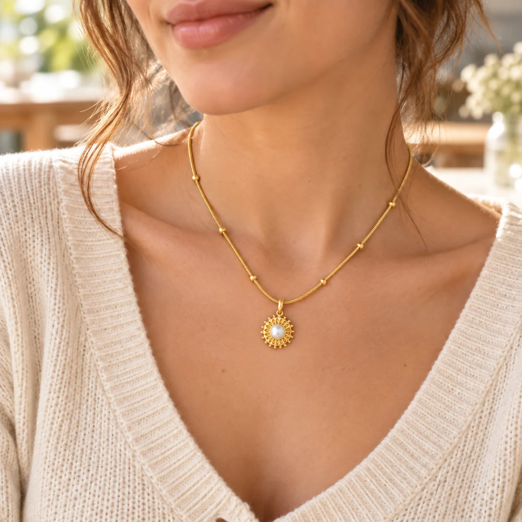 A close-up lifestyle shot of a woman wearing a delicate gold chain necklace with a sunflower pendant featuring a white pearl center, styled with a cream-colored knit sweater.